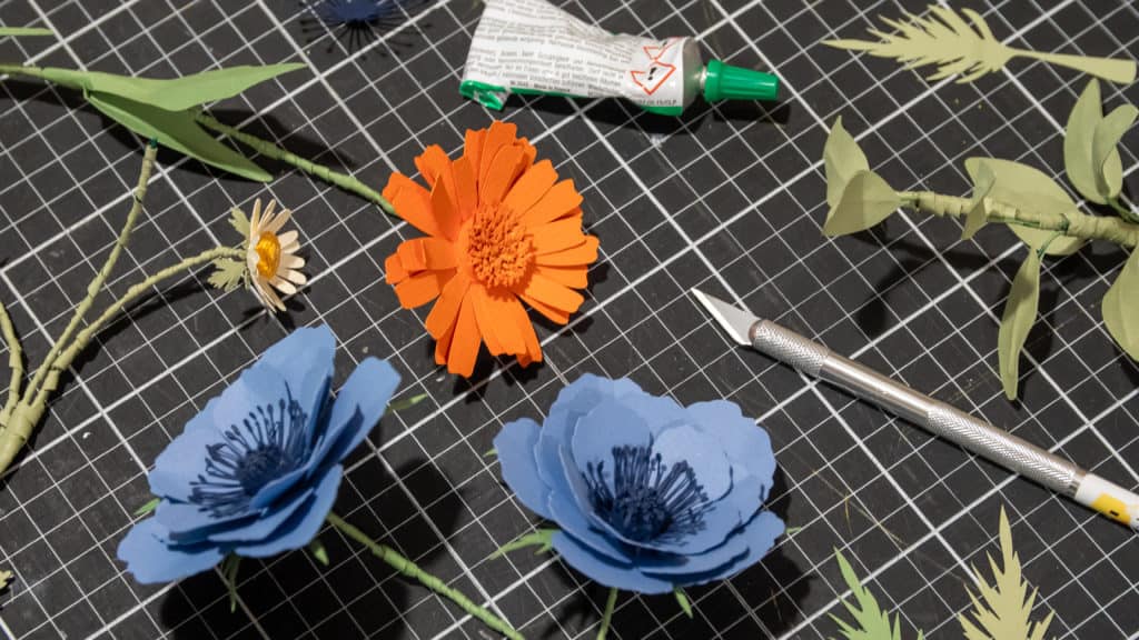 Fleurs en papier, scalpel et colle sur un tapis de découpe, fabrication du décor du spot Apilis