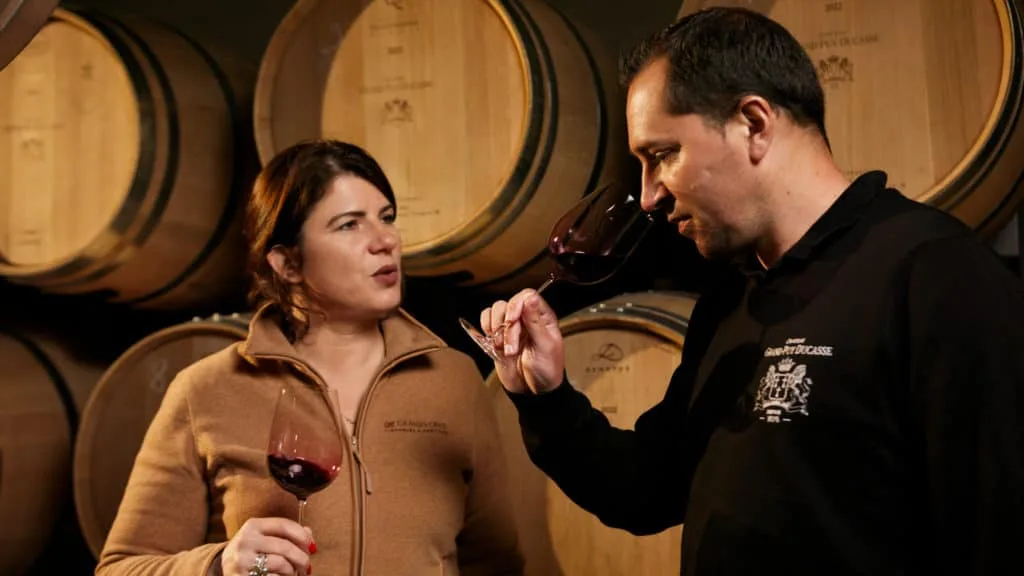 Dégustation au chai : deux membres de l’équipe du château au verre devant les barriques du millésime