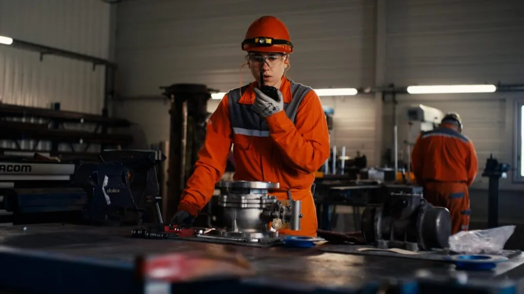 Technicienne de maintenance en combinaison orange à la radio devant une pièce démontée, EcoTitanium